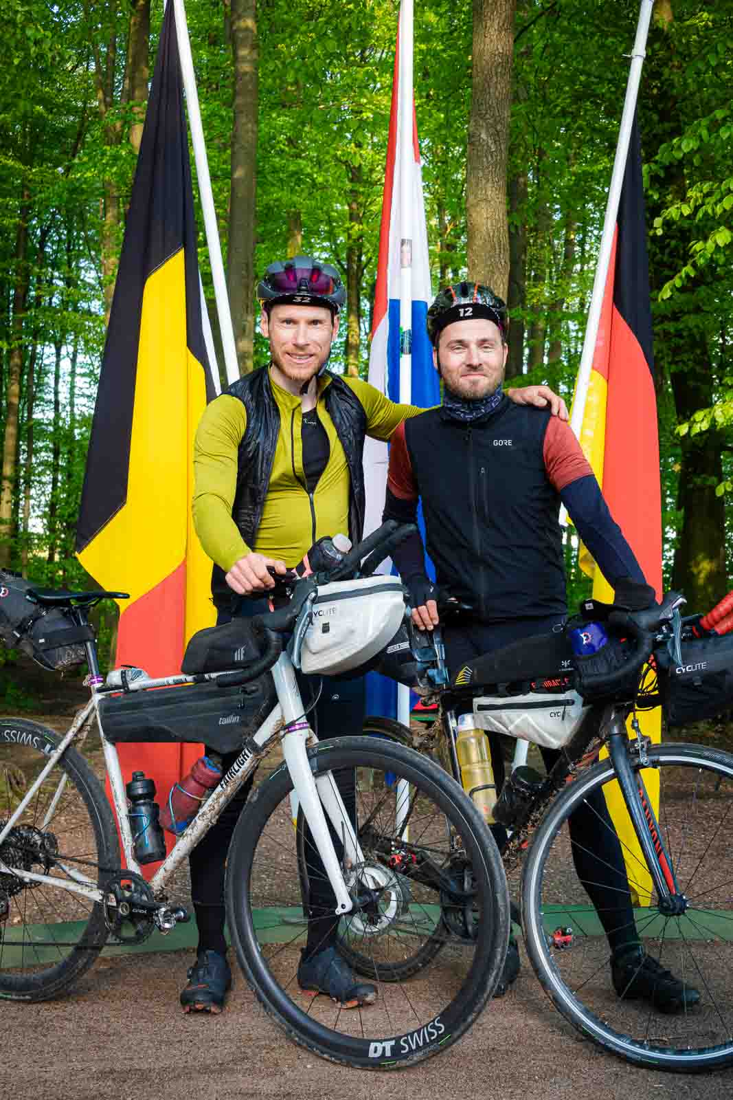 Des cyclistes se tiennent devant trois drapeaux au tripoint Au tripoint de l'Allemagne, de la Belgique et des Pays-Bas, deux cyclistes se tiennent et rient vers la caméra.