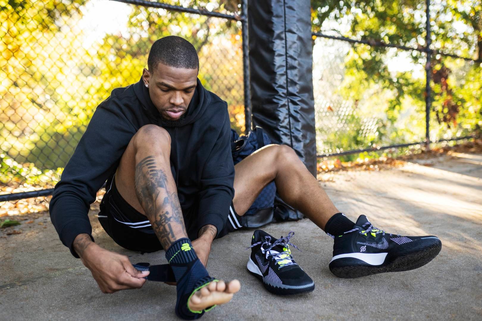 Basketballspieler mit Hoodie zieht sich am Rand eines Streetball-Courts eine Sprunggelenkbandage mit Gurt an