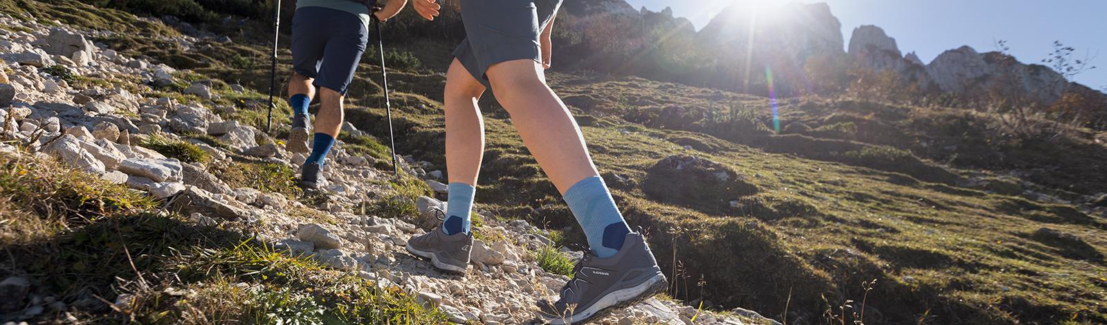 Pas sûr en terrain difficile : Nos chaussettes Outdoor Performance Mid Cut Gros plan sur le bas des jambes d'une randonneuse en chaussures de randonnée et avec des chaussettes de randonnée mi-longues vertes qui monte un chemin rocailleux, en arrière-plan son partenaire de randonnée