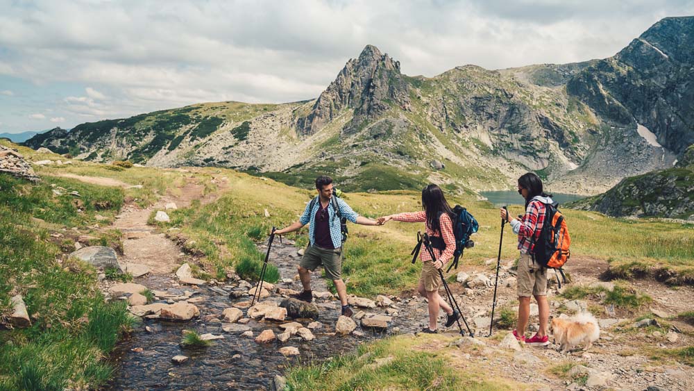 Gemeinsam über Stock und Stein Drei Wanderer, zwei Frauen und ein Mann, überqueren gemeinsam einen Gebirgsbach in alpiner Landschaft mit felsigen Gipfeln im Hintergrund. Der Mann reicht einer Frau die Hand zur Unterstützung