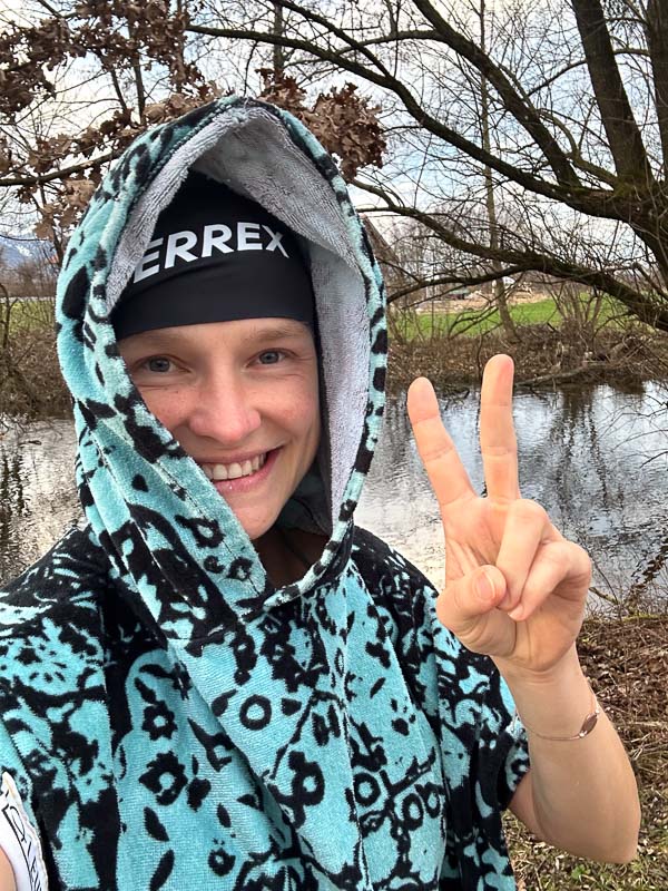 Anna Hahner nach dem Eisbaden mit Terrex-Beanie Anna Hahner im Terrex-Beanie und buntem Poncho, Handtuch um die Schultern gewickelt, zeigt das Friedenszeichen.