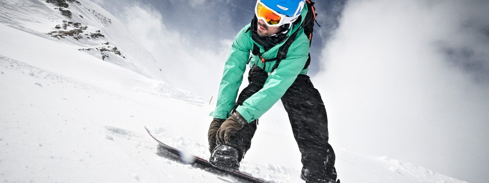 Sitzt alles richtig? Dann genieß' die Abfahrt mit uns! Leichte Froschperspektive: Snowboarder in grüner Jacke befestigt mit seinen Händen eine Bindung und blickt nach unten während um ihn herum viel Schnee aufgewirbelt wird