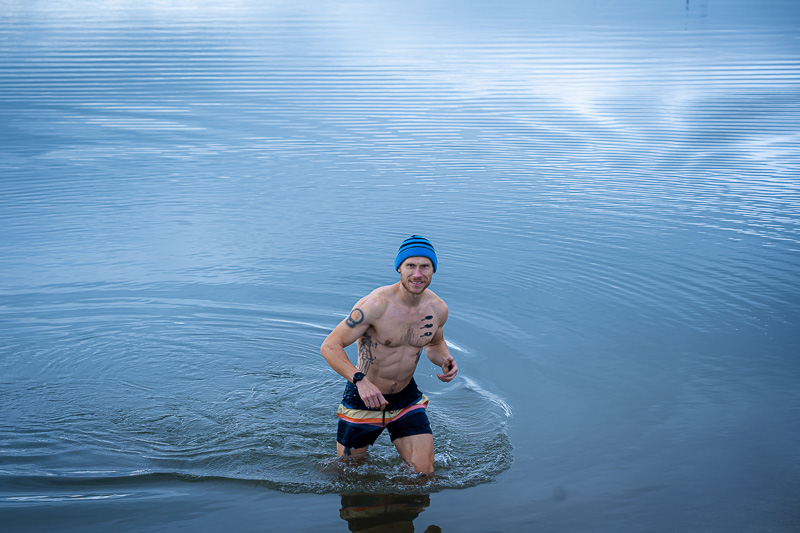 Schnell raus aus dem Eisbad! Ein Mann watet aus dem See, während er beim Eisbaden in Badehose und mit blauer Mütze in der herbstlichen Umgebung fotografiert wird.