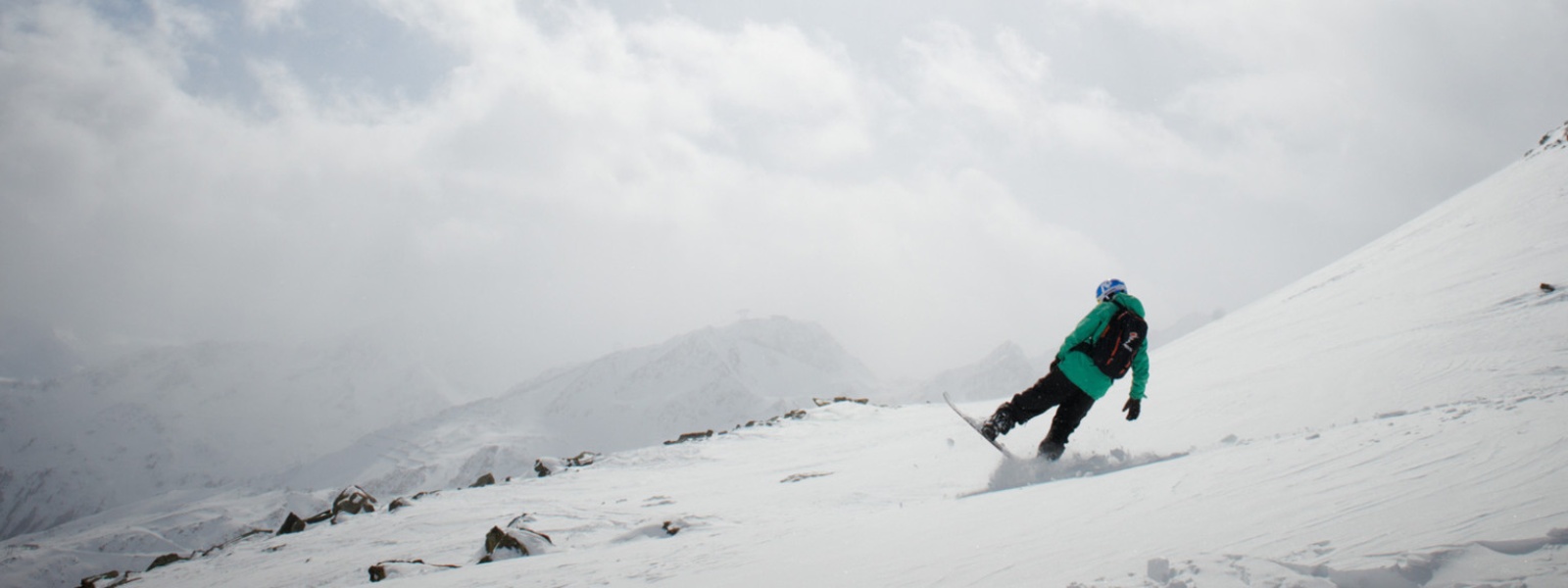 Comment préparer ton corps pour les prochaines descentes spectaculaires, tu le découvriras ici avec nous Snowboarder en veste verte freine avec l'arrière de sa planche devant une bosse, en arrière-plan on voit des sommets enneigés à travers les nuages