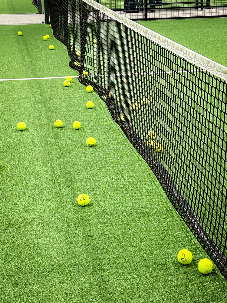Der Platz erzählt die Geschichte eines intensiven Padel-Spiels. Mehrere gelbe Padel-Bälle liegen verstreut auf einem grünen Kunstrasenplatz direkt neben dem Netz – ein lebendiger Moment nach intensiven Ballwechseln.