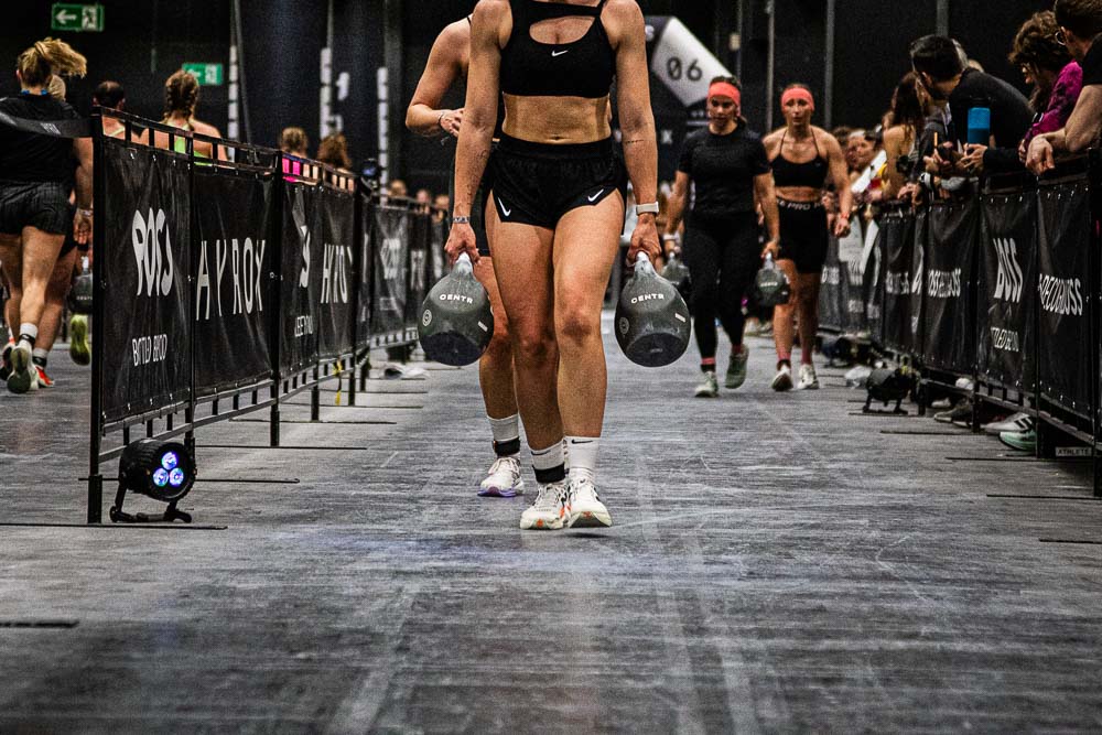Athletin trägt beim HYROX-Wettkampf zwei schwere Kettlebells über die Laufstrecke, während weitere Teilnehmende hinter ihr folgen und Zuschauerinnen und Zuschauer die Strecke säumen; die Szene zeigt Griffkraft