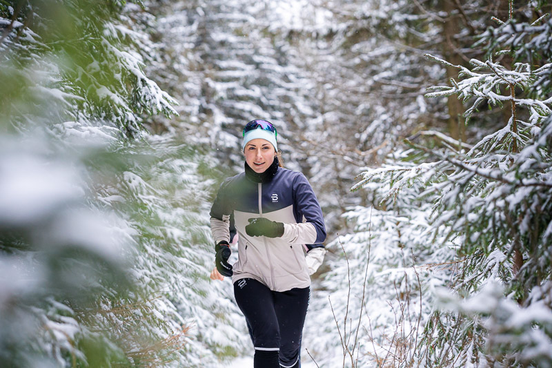 Winterlauf: Zwischen verschneiten Tannen auf dem Pfad der Entschlossenheit. Eine Läuferin joggt durch einen verschneiten Waldweg. Die Bäume um sie herum sind dicht mit Schnee bedeckt. Sie trägt eine hellgraue Laufjacke mit schwarzen Akzenten, eine schwarze Hose und eine blaue Stirnbinde mit Sonnenbrille.