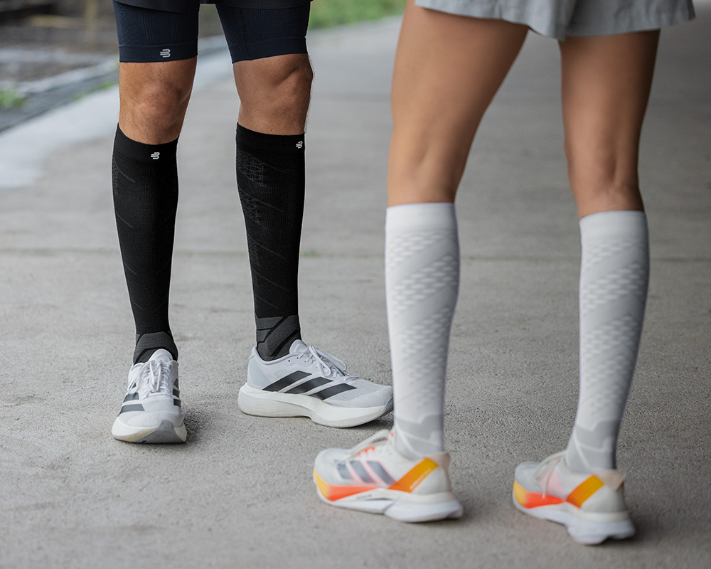 Parfaites pour courir en ville : nos Run Performance Compression Socks Vue en contre-plongée : des coureuses avec des chaussettes de course violettes et blanches courent à travers une ville animée, en arrière-plan des gens, des maisons et des lampadaires