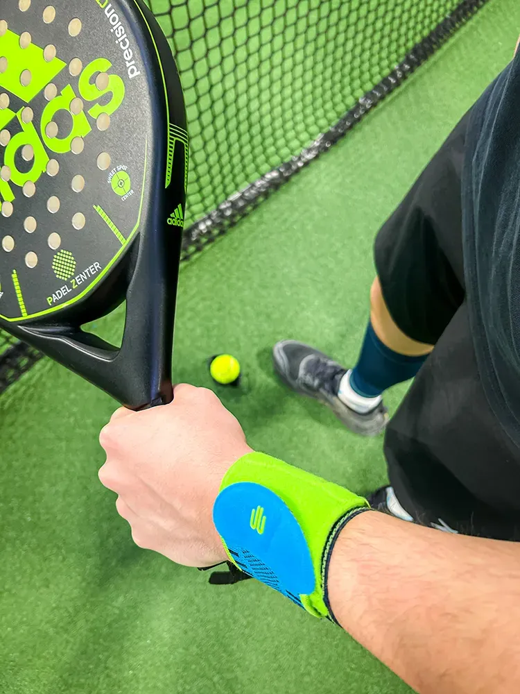 Konzentration am Netz: Bereit für den nächsten Schlag. Nahaufnahme eines Spielers auf einem Padelplatz, der einen Adidas-Padelschläger hält. Der Sportler trägt eine neonfarbene Handgelenkbandage, während ein Ball am Boden liegt.