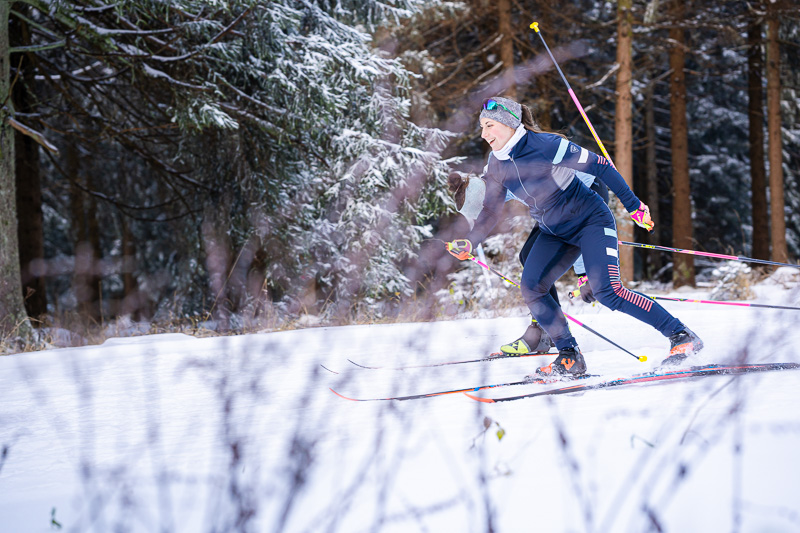 Dynamik und Präzision beim Langlauf durch den verschneiten Wald. Eine Sportlerin beim Langlaufen in einem verschneiten Wald. Sie bewegt sich energisch durch den tiefen Schnee, mit professioneller Langlaufausrüstung und moderner Sportkleidung.