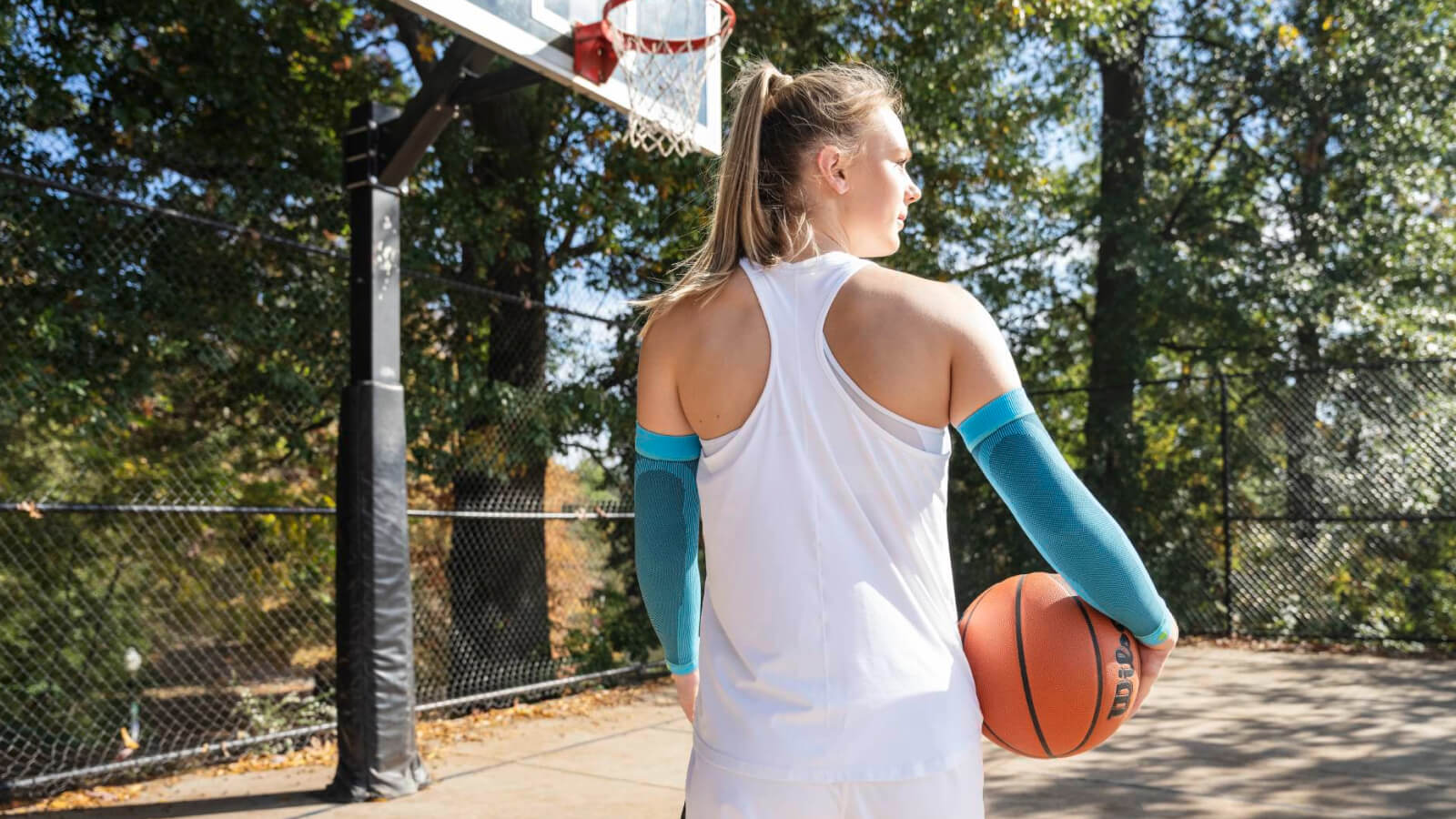 Eine Basketballspielerin steht auf einem Outdoor-Platz mit einem Basketball in der rechten Hand. Sie trägt hellblaue Kompressions-Arm Sleeves und blickt konzentriert auf den Korb.