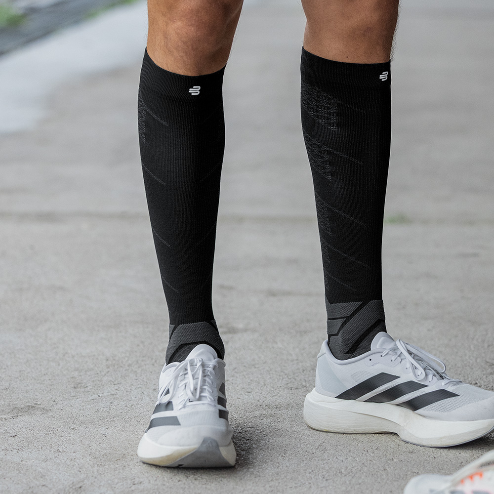 Stabiler Stand mit starker Kompression: Die schwarzen Run Performance Socks im Fokus. Ein sportlicher Mann steht frontal auf Asphalt, bekleidet mit schwarzen Kompressionsstrümpfen von Bauerfeind und weißen Laufschuhen. Die geradlinige Haltung und die Muskelspannung in den Waden betonen den stabilisierenden Effekt der Socken im Stehen.