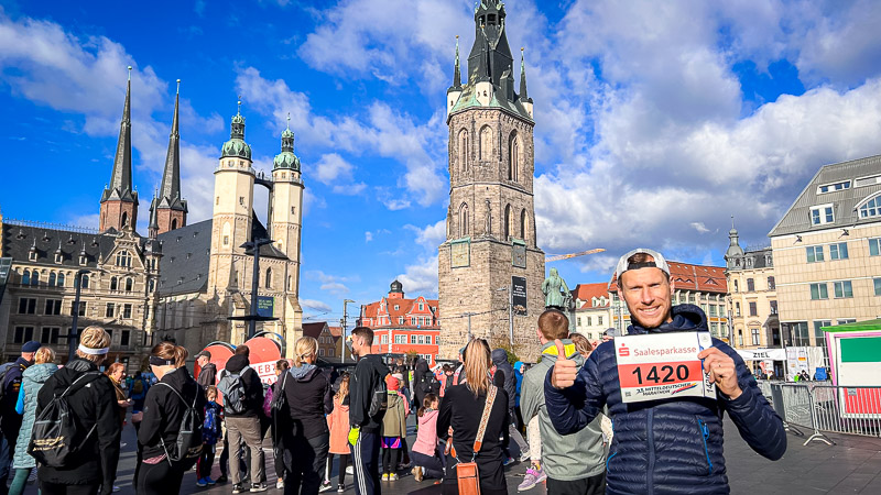 Teilnehmer am Startpunkt des Mitteldeutschen Halbmarathons auf dem Marktplatz in Halle (Saale) vor der Kulisse der Marktkirche und dem Roten Turm. Ein Teilnehmer des Mitteldeutschen Halbmarathons steht auf dem Marktplatz in Halle und hält lächelnd seine Startnummer hoch. Als Zeichen seines Enthusiasmus zeigt der den Daumen nach oben. Er ist umgeben von Zuschauern und verschiedenen Bauwerken.
