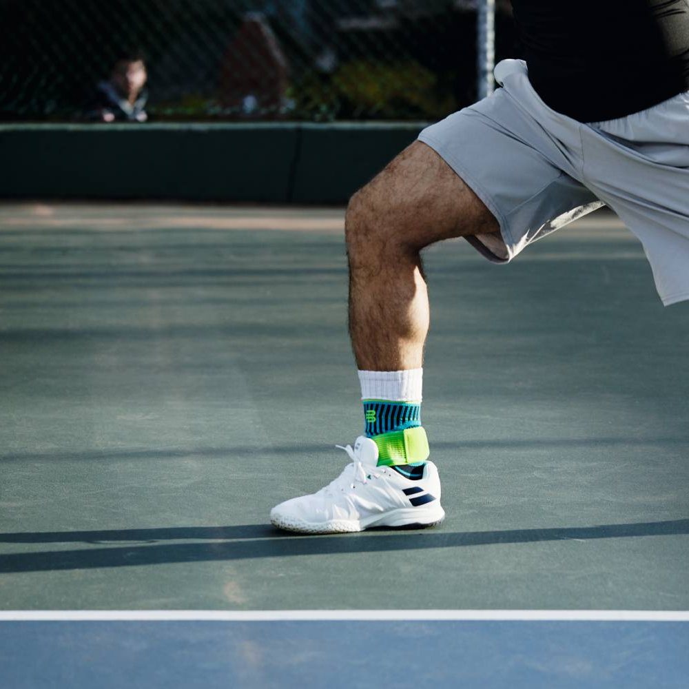 Jeder Schritt zählt – Unterstützung für schnelle Richtungswechsel! Ein Tennisspieler auf dem Hartplatz, in Bewegung beim Aufschlag. Er trägt die Bauerfeind Sports Ankle Support Rivera in Blau-Grün über seinen weißen Sportsocken.