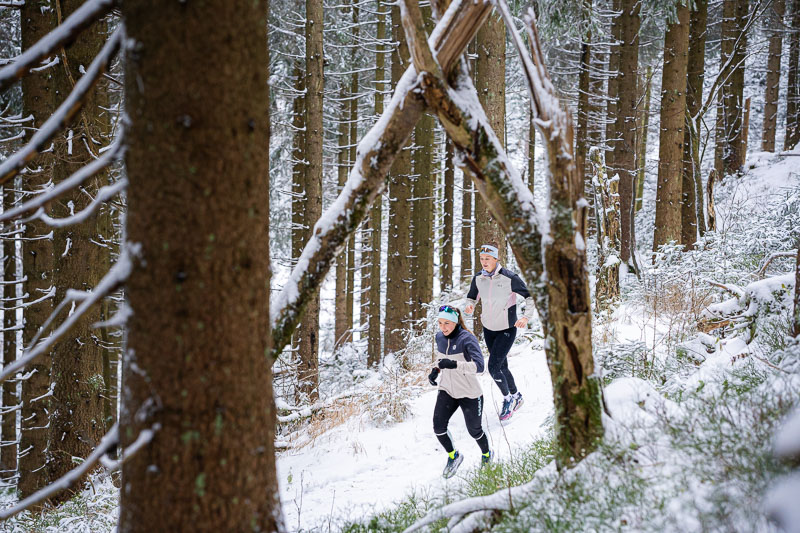 Winterlauf unter dem Torbogen der Natur Zwei Läufer joggen durch einen verschneiten Wald. Der Weg ist umgeben von hohen Tannenbäumen, die mit Schnee bedeckt sind, während ein natürlicher Torbogen aus Ästen den Pfad überspannt.