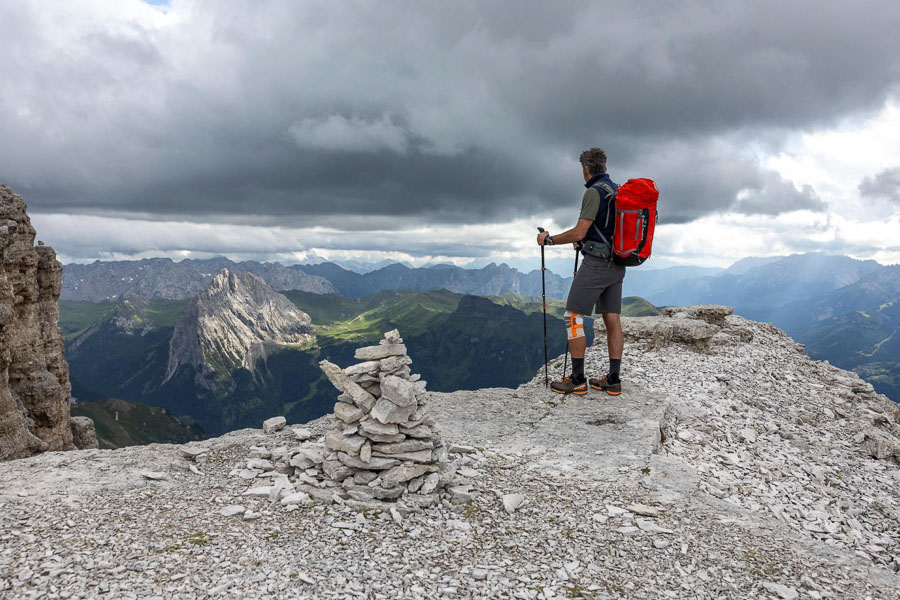 Randonneur sur un promontoire rocheux avec un genouillère de randonnée Une personne avec un sac à dos rouge se tient sur un promontoire rocheux dans les Dolomites et regarde au loin. Le ciel est nuageux et il y a une construction en cairn au premier plan.