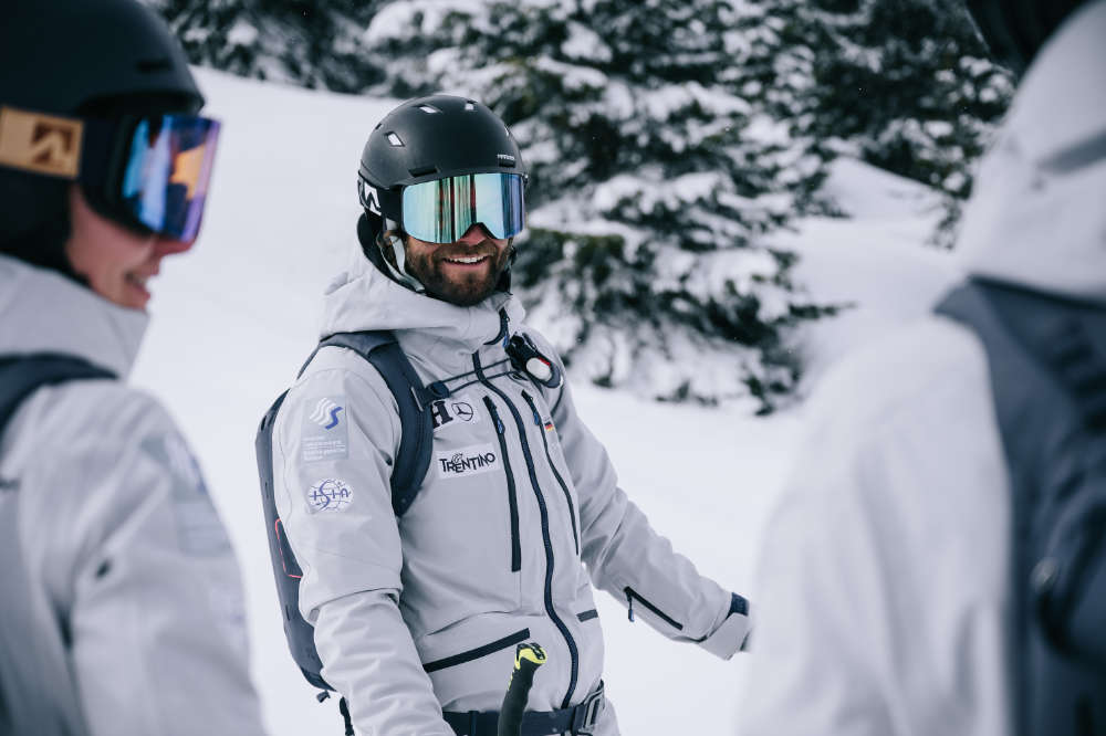 Skilehrer aus unserem Sportpartnerverband DSLV Gut gelaunter Skifahrer mit schwarzem Helm	 grauer Jacke und Rucksack	 Aufnahme zwischen zwei weiteren Skifahrern