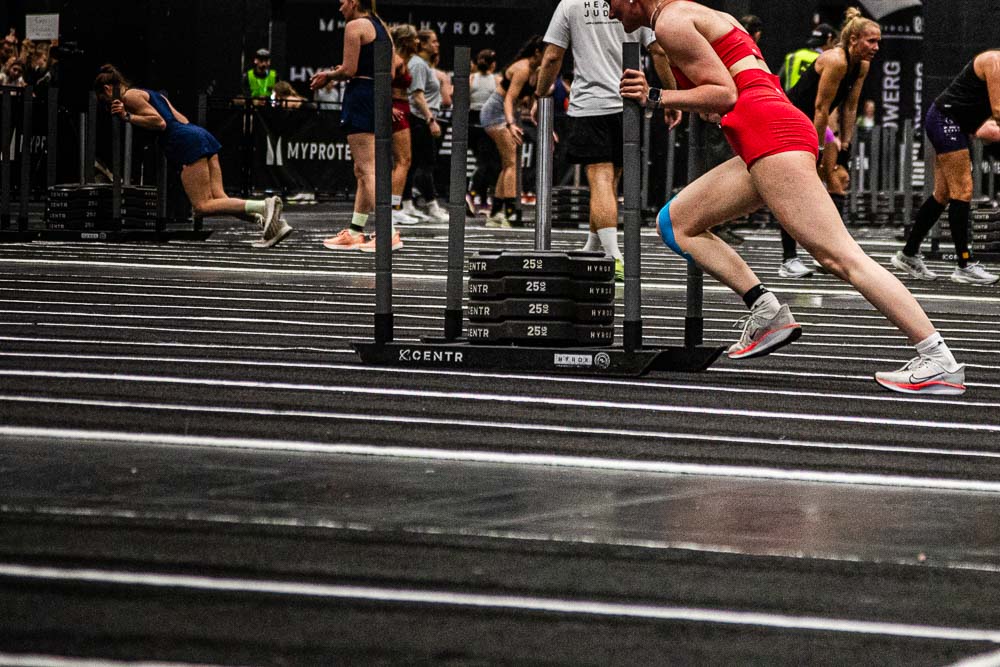 Athletin in rotem Sportoutfit schiebt bei einem HYROX-Wettkampf einen schweren Trainingsschlitten über die markierte Indoor-Fläche, während weitere Teilnehmende im Hintergrund an ihren Stationen arbeiten