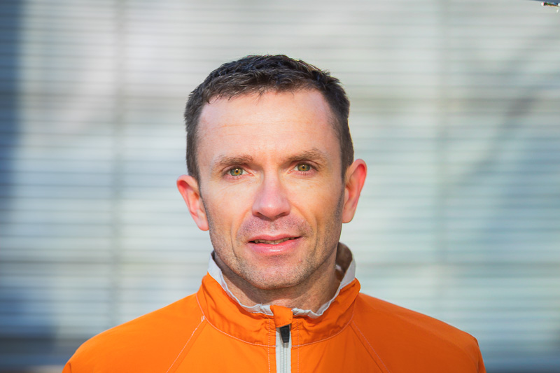 Ein Portrait in der Sonne: Christian Seiler Ein Mann in einer leuchtend orangefarbenen Jacke blickt lächelnd in die Kamera. Der Hintergrund zeigt ein modernes Gebäude mit Bäumen und blühenden Pflanzen.