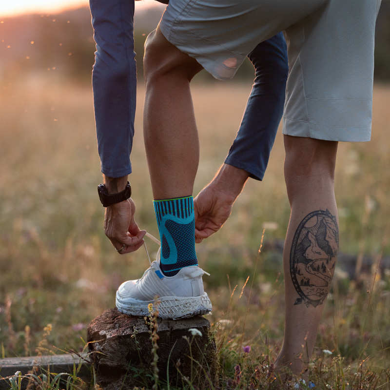 Sicherheit und Entlastung beim Laufen durch die Bauerfeind Sports Achilles Support Mann mit Fußbandage bindet Laufschuh mit seinem Fuß auf einem Baumstumpf
