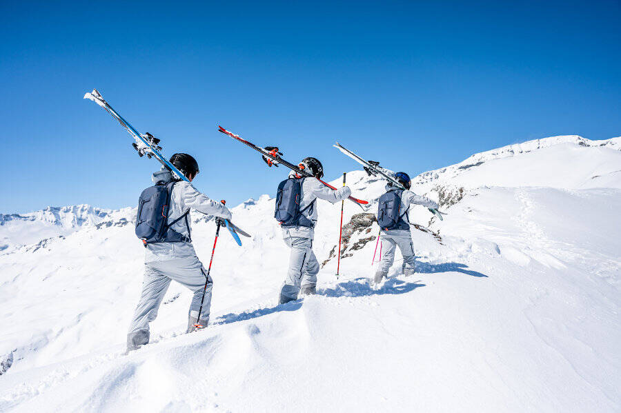 Prépare tes skis et accompagne-nous sur la montagne Trois skieurs portent leurs skis sur les épaules à travers une neige profonde en montant une montagne
