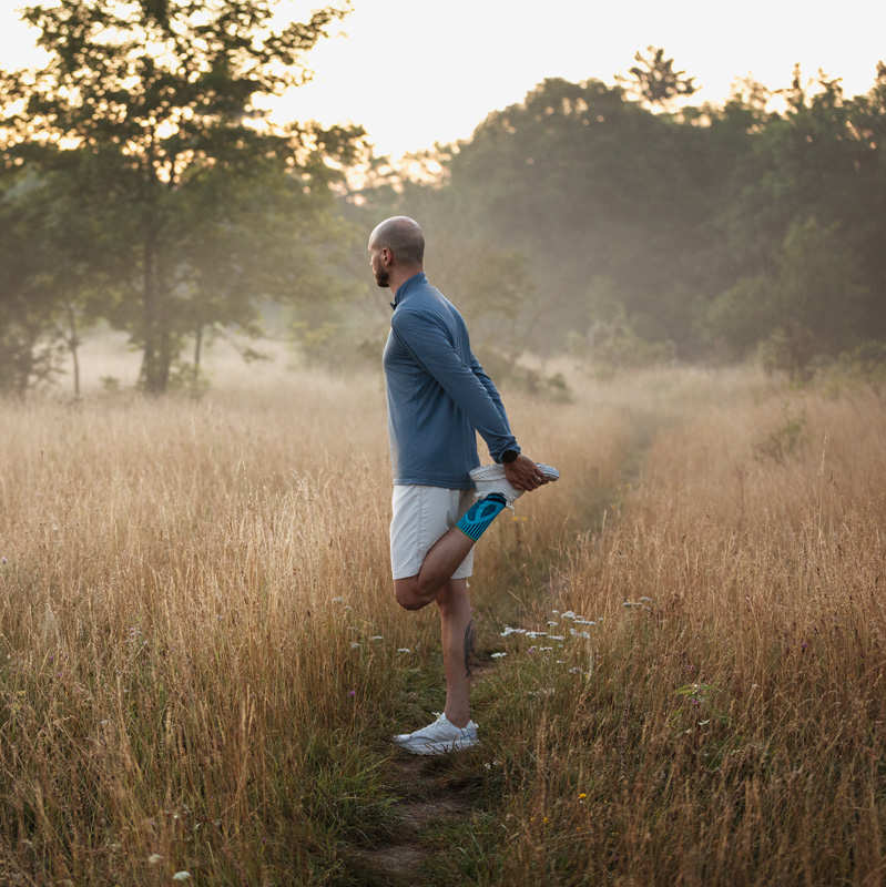 Entgehe Schmerzen beim Laufen durch die Bauerfeind Sports Achilles Support Mann dehnt sich beim Sonnenaufgang auf einer Wiese mit hohem Gras